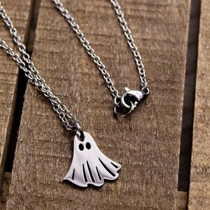 May include: A silver-colored necklace with a ghost pendant. The ghost pendant has two black eye holes and a flowing, sheet-like body. The chain is a delicate, linked design. The necklace is displayed on a wooden surface.