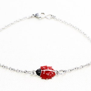 Puede incluir: Un brazalete de cadena de plata con un dije de mariquita roja. La mariquita tiene manchas negras y una cabeza negra.