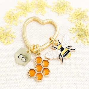 May include: Gold heart-shaped keychain with three charms: a bee with black and yellow stripes, a honeycomb charm, and a hexagonal charm with the inscription "C♡".
