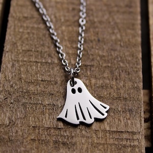 May include: A silver-colored necklace with a ghost pendant. The ghost pendant has two black eyes and a cutout design for the mouth and bottom. The necklace is displayed on a wooden surface.