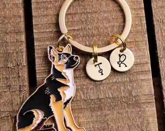 Könnte beinhalten: Ein goldfarbener Schlüsselanhänger mit einem Schäferhund-Anhänger mit schwarzen, braunen und weißen Emaille-Details. Zwei kleine, runde Anhänger mit den Buchstaben "T" und "R" hängen am Schlüsselanhänger.