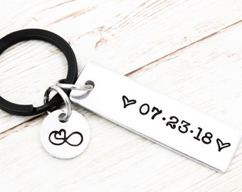 May include: A silver-colored rectangular keychain with the date "07.23.18" and two hearts stamped on it. A small round charm with an infinity symbol is attached. The keychain has a black ring.