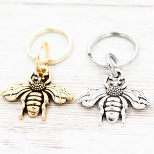 Puede incluir: Dos llaveros con forma de abeja, uno dorado y otro plateado, sobre un fondo blanco.