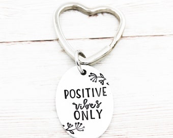 Puede incluir: Llavero de corazón plateado con un dije ovalado que dice "POSITIVE vibes ONLY" en letras negras con detalles florales.
