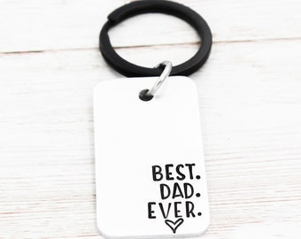 May include: A white metal keychain with a black key ring. The keychain has the text "BEST. DAD. EVER." engraved on it with a small heart symbol.