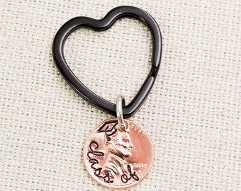 Può includere: Un portachiavi a forma di cuore nero con una moneta di rame attaccata. La moneta ha le parole "Class of" incise su di essa con un cappello di laurea sopra le parole.