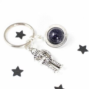 May include: A silver astronaut keychain with a black galaxy charm and a silver key ring. The keychain is surrounded by black paper stars on a white background.