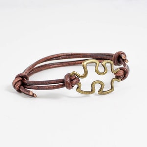 May include: A brown leather bracelet with a gold-toned puzzle piece charm.