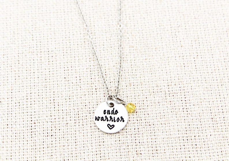 May include: Silver necklace with a round pendant that says "endo warrior" with a small heart below the text. A small yellow bead hangs from the pendant.