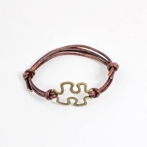 May include: A brown leather bracelet with a gold-toned puzzle piece charm.