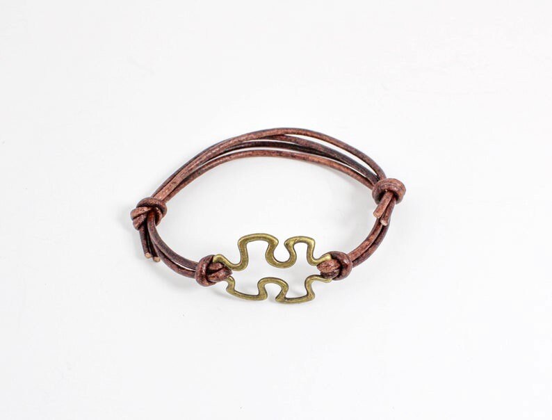May include: A brown leather bracelet with a gold-toned puzzle piece charm.