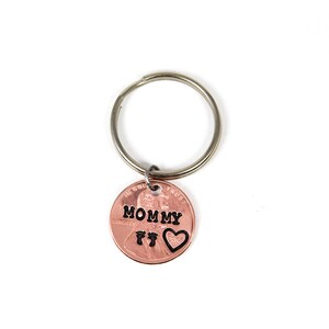 Puede incluir: Un llavero de un centavo de cobre con las palabras "Mommy" y dos huellas de pies, y un coraz&oacute;n. El llavero est&aacute; unido a un llavero de plata.