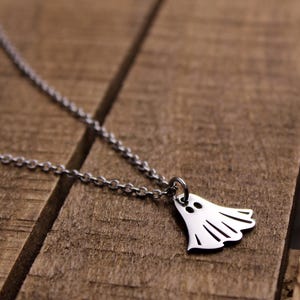 May include: A silver-colored necklace featuring a ghost pendant. The ghost has two eye holes and a curved bottom with vertical lines. The necklace chain is a delicate link style. The pendant is approximately 0.75 inches wide.