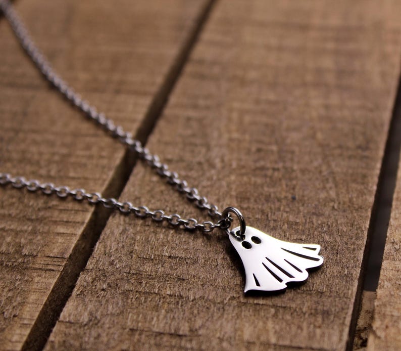 May include: A silver-colored necklace featuring a ghost pendant. The ghost has two eye holes and a curved bottom with vertical lines. The necklace chain is a delicate link style. The pendant is approximately 0.75 inches wide.