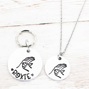 May include: A silver dog tag with a handprint design and the name "ROXIE" engraved on it. A matching silver necklace with the same handprint design is also shown.