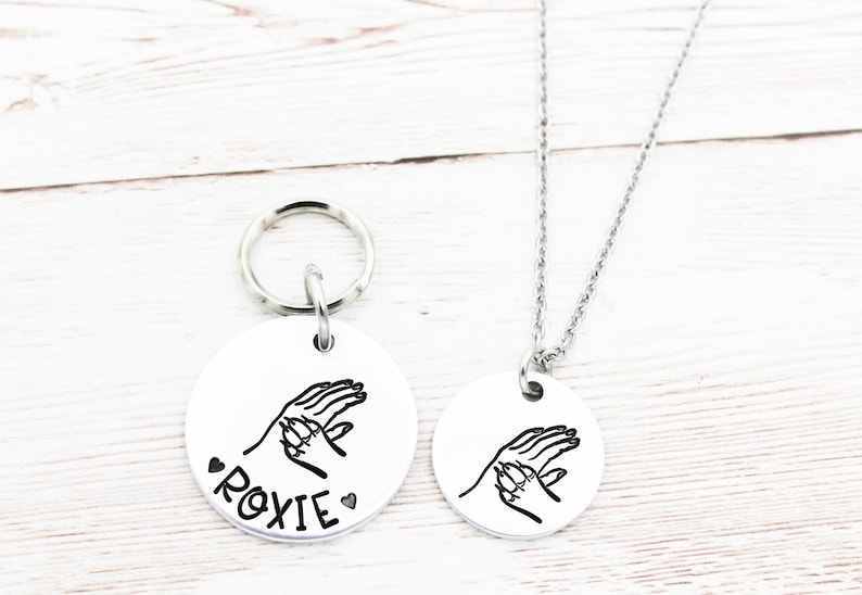 May include: A silver dog tag with a handprint design and the name "ROXIE" engraved on it. A matching silver necklace with the same handprint design is also shown.