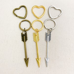Op de afbeelding: Drie sleutelhangers met pijl-bedels. De bedels zijn zilver, goud en brons. De sleutelhangers zijn bevestigd aan hartvormige ringen.