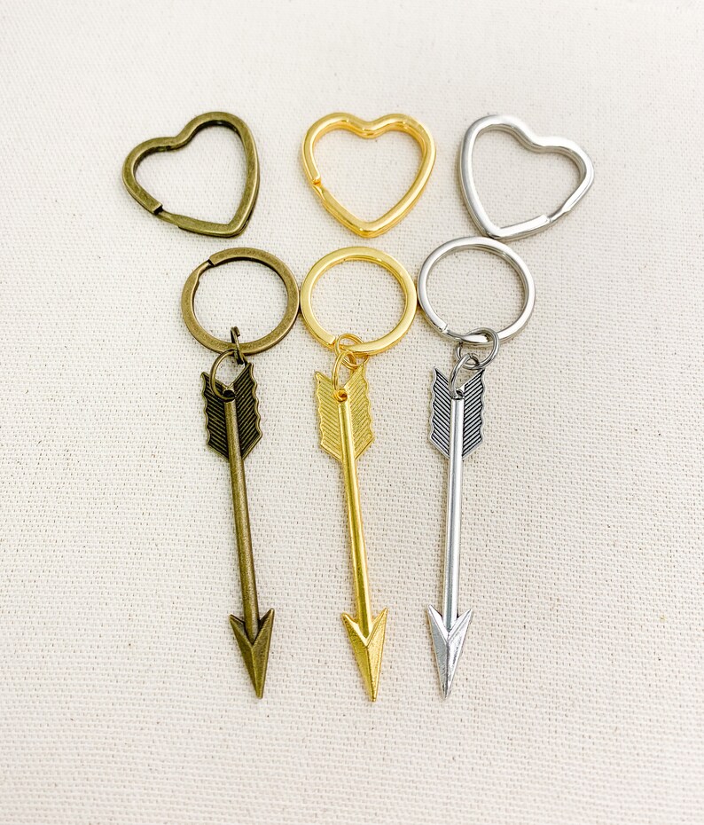 Op de afbeelding: Drie sleutelhangers met pijl-bedels. De bedels zijn zilver, goud en brons. De sleutelhangers zijn bevestigd aan hartvormige ringen.