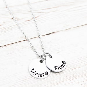 May include: Silver necklace with two round pendants. The pendants are engraved with the names "Walter" and "Pippi" and a paw print.