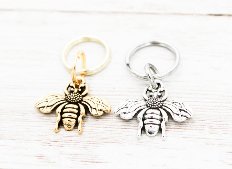 Puede incluir: Dos llaveros con forma de abeja, uno dorado y otro plateado, sobre un fondo blanco.