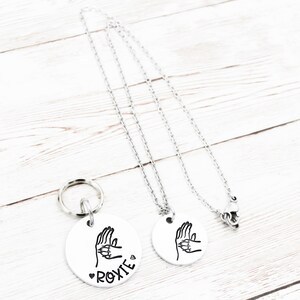 May include: Two silver necklaces with round pendants. The pendants feature a hand sign with the word "ROXIE" on one pendant.