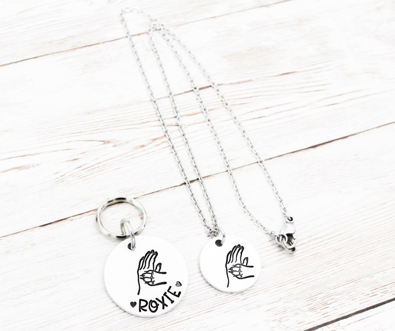 May include: Two silver necklaces with round pendants. The pendants feature a hand sign with the word "ROXIE" on one pendant.