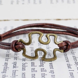 May include: A brown leather bracelet with a brass puzzle piece charm. The bracelet is adjustable and has a simple design.