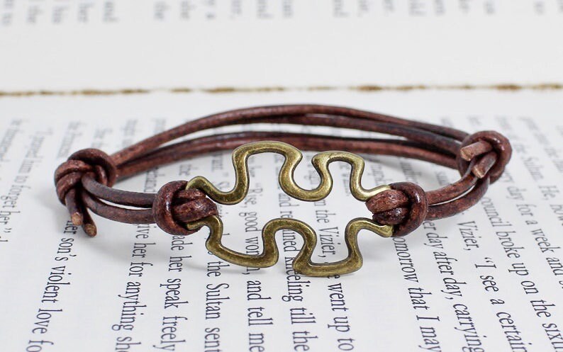 May include: A brown leather bracelet with a brass puzzle piece charm. The bracelet is adjustable and has a simple design.