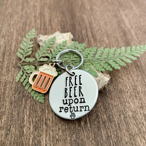 K&ouml;nnte beinhalten: Ein silberner Schl&uuml;sselanh&auml;nger mit einem Bierkrug-Anh&auml;nger und dem Text "FREE BEER upon return" mit einem Pfotenabdruck unter dem Text.