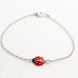 Puede incluir: Un brazalete de cadena plateada con un dije de mariquita roja. La mariquita tiene manchas negras y una cabeza negra.