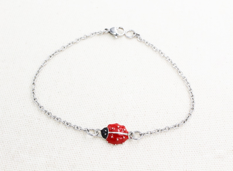Puede incluir: Un brazalete de cadena plateada con un dije de mariquita roja. La mariquita tiene manchas negras y una cabeza negra.