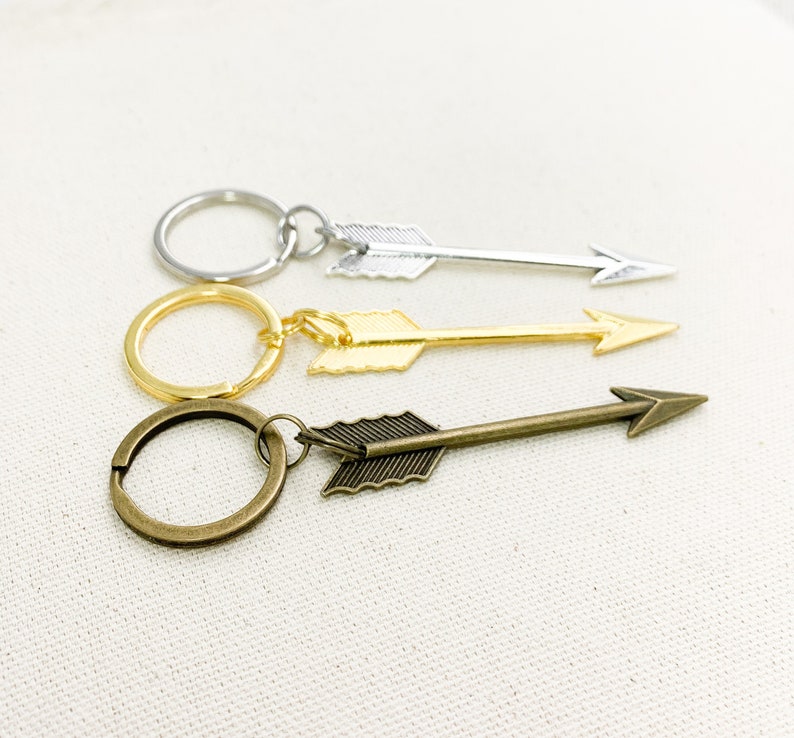 Op de afbeelding: Drie pijlvormige sleutelhangers met verschillende afwerkingen: zilver, goud en brons. Elke sleutelhanger heeft een ring om sleutels aan te bevestigen.