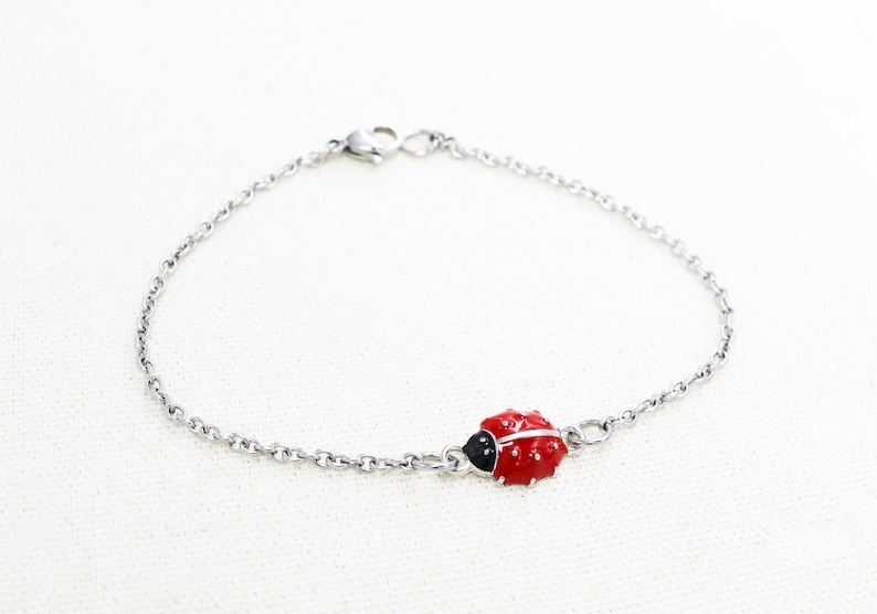 Puede incluir: Un brazalete de cadena de plata con un dije de mariquita roja. La mariquita tiene manchas negras y una cabeza negra.