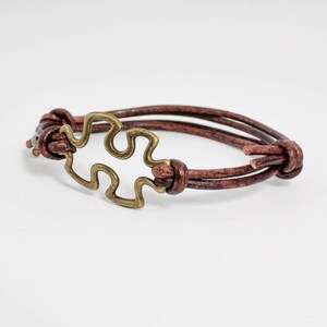 May include: A brown leather bracelet with a gold-toned puzzle piece charm.