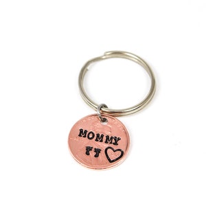Puede incluir: Llavero de un centavo de cobre con un llavero de plata. El centavo tiene las palabras "MOMMY" y dos huellas de pies con un coraz&oacute;n grabado.