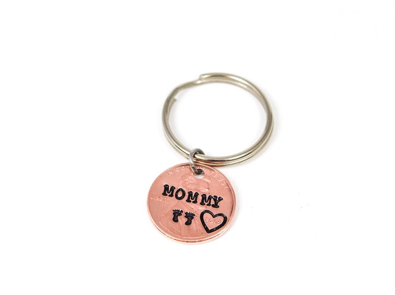 Puede incluir: Llavero de un centavo de cobre con un llavero de plata. El centavo tiene las palabras "MOMMY" y dos huellas de pies con un coraz&oacute;n grabado.