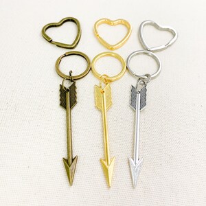 Op de afbeelding: Drie pijl sleutelhangers met verschillende afwerkingen: brons, goud en zilver. Elke sleutelhanger heeft een hartvormige ring en een kleine pijl hanger.