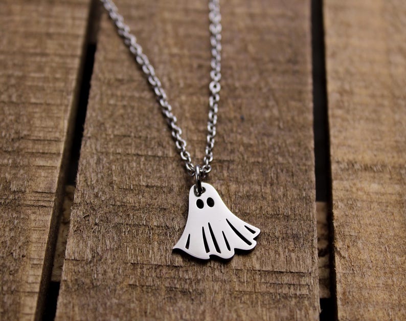 May include: A silver-colored necklace with a ghost pendant. The ghost pendant has two black eyes and a cutout design for the mouth and bottom. The necklace is displayed on a wooden surface.