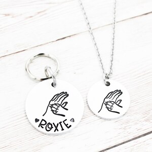 May include: A silver dog tag with a paw print design and the name "ROXIE" engraved on it. The tag is attached to a silver key ring. A matching silver necklace with the same paw print design is also shown.