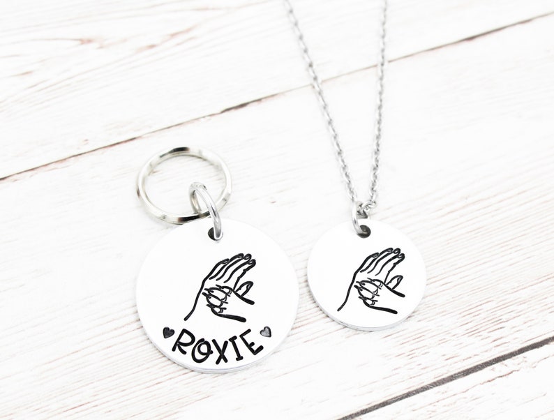 May include: A silver dog tag with a paw print design and the name "ROXIE" engraved on it. The tag is attached to a silver key ring. A matching silver necklace with the same paw print design is also shown.
