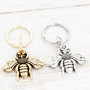 Puede incluir: Dos llaveros con forma de abeja, uno dorado y otro plateado, con un dise&ntilde;o de anillo simple. Los llaveros est&aacute;n sobre una superficie de madera blanca.