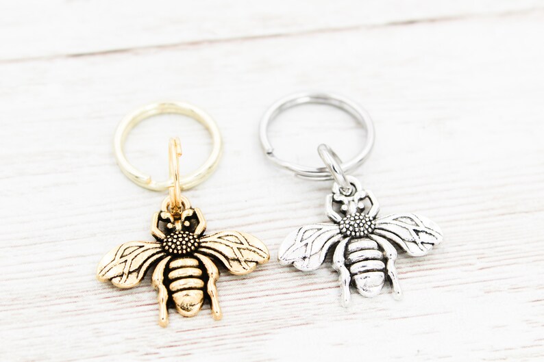 Puede incluir: Dos llaveros con forma de abeja, uno dorado y otro plateado, con un dise&ntilde;o de anillo simple. Los llaveros est&aacute;n sobre una superficie de madera blanca.