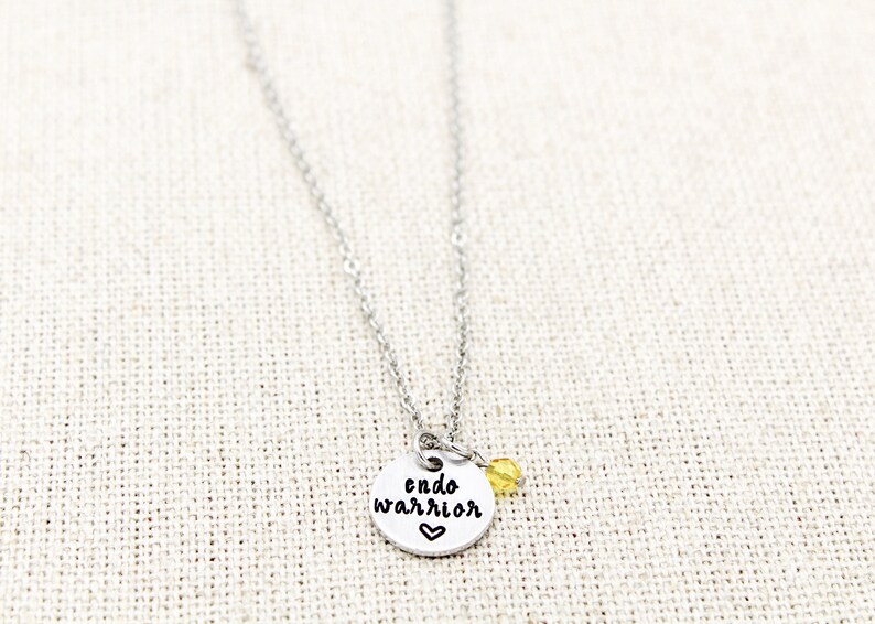May include: Silver necklace with a round pendant that says "endo warrior" with a small heart below the text. A small yellow bead hangs from the pendant.