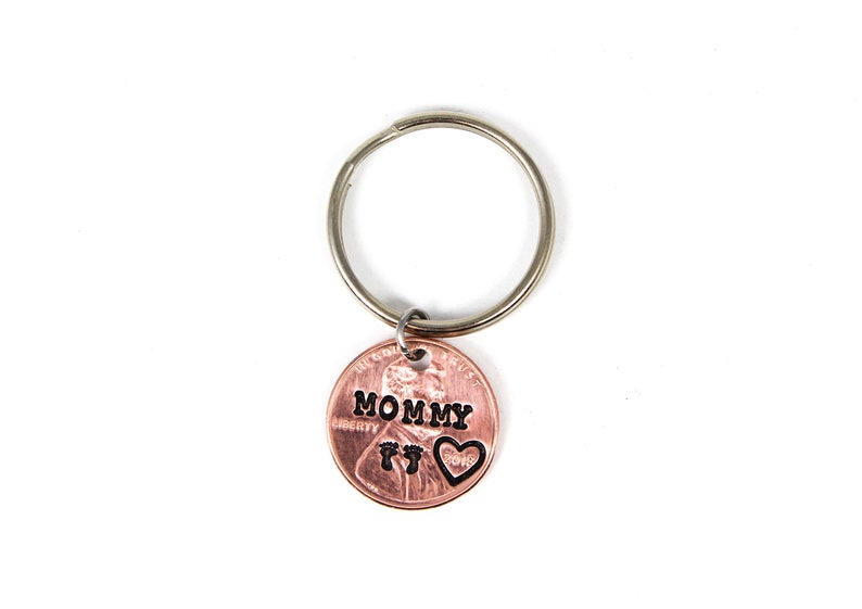 Puede incluir: Un llavero de un centavo de cobre con las palabras "Mommy" y un coraz&oacute;n con huellas de pies grabadas.