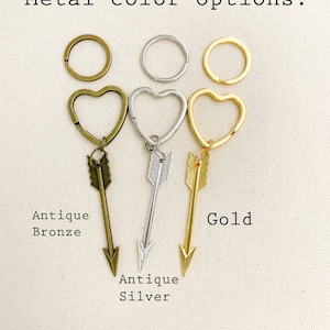 Op de afbeelding: Drie sleutelhangers met hartvormige ringen en pijlbedels. De sleutelhangers zijn in antiek brons, antiek zilver en goud.