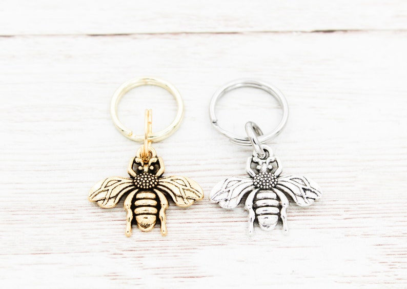 Puede incluir: Dos abalorios de llavero con forma de abeja, uno dorado y otro plateado, en llaveros plateados. Los abalorios de abeja est&aacute;n detallados con alas, un cuerpo y un aguij&oacute;n.