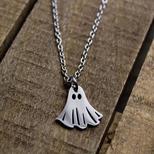 May include: A silver-colored ghost pendant necklace. The pendant is a stylized ghost shape with two eye holes and a wavy bottom edge. The necklace chain is also silver and is resting on a wooden surface.