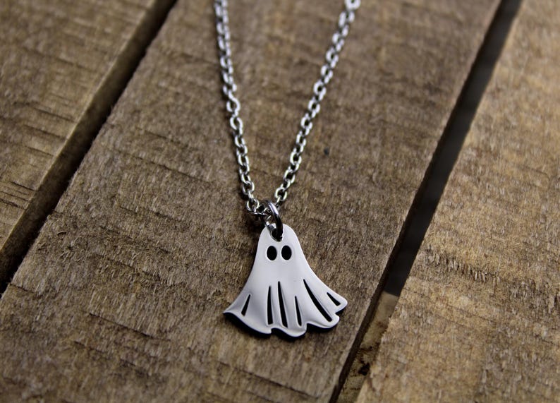 May include: A silver-colored ghost pendant necklace. The pendant is a stylized ghost shape with two eye holes and a wavy bottom edge. The necklace chain is also silver and is resting on a wooden surface.