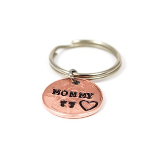Puede incluir: Un llavero de un centavo de cobre con las palabras "Mommy" y un coraz&oacute;n con huellas de pies grabadas.