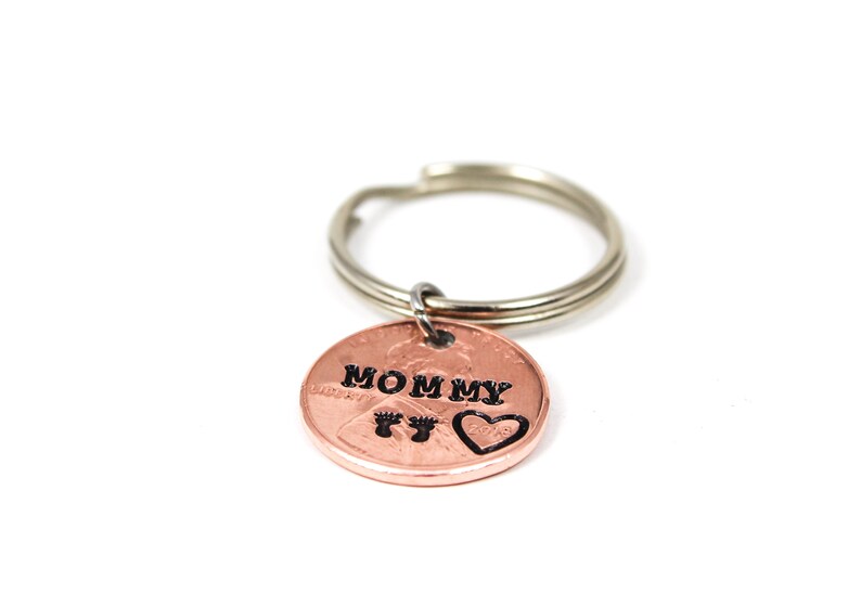 Puede incluir: Un llavero de un centavo de cobre con las palabras "Mommy" y un coraz&oacute;n con huellas de pies grabadas.